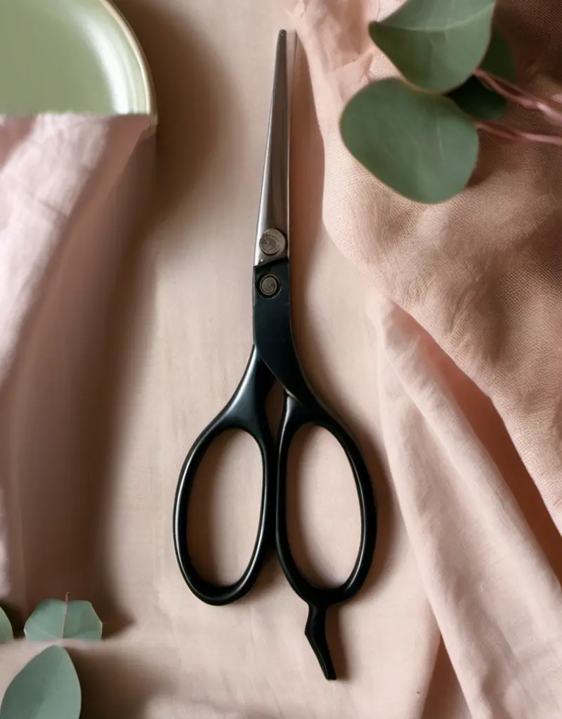 Black hairdressing shears on blush linen beside a celadon dish and eucalyptus leaves